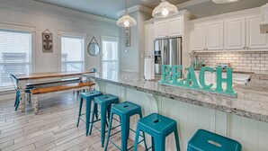 Une cuisine avec des meubles blancs, un réfrigérateur en acier inoxydable, des tabourets de bar turquoise et une pancarte « PLAGE » posée sur le comptoir.