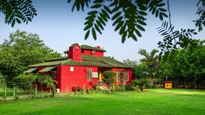 A red house with a green roof, surrounded by a well-maintained lawn and trees. - Organic Farm with kitchen, AC rooms, Pet-friendly 7 Kms from the city (Jhansi)