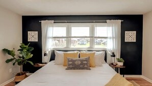 Une chambre à coucher avec un grand lit, deux tables de chevet, une fenêtre avec des rideaux et des panneaux décoratifs sur les murs.