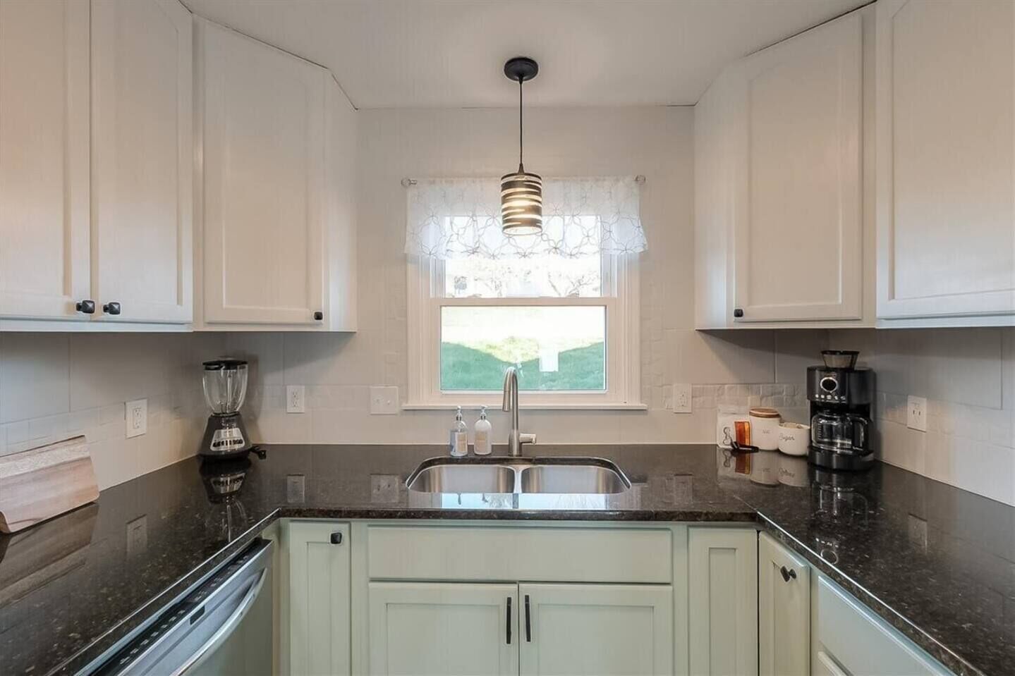 A kitchen with white cabinets, a double sink, a blender, a coffee maker, and a window with a view of greenery.
