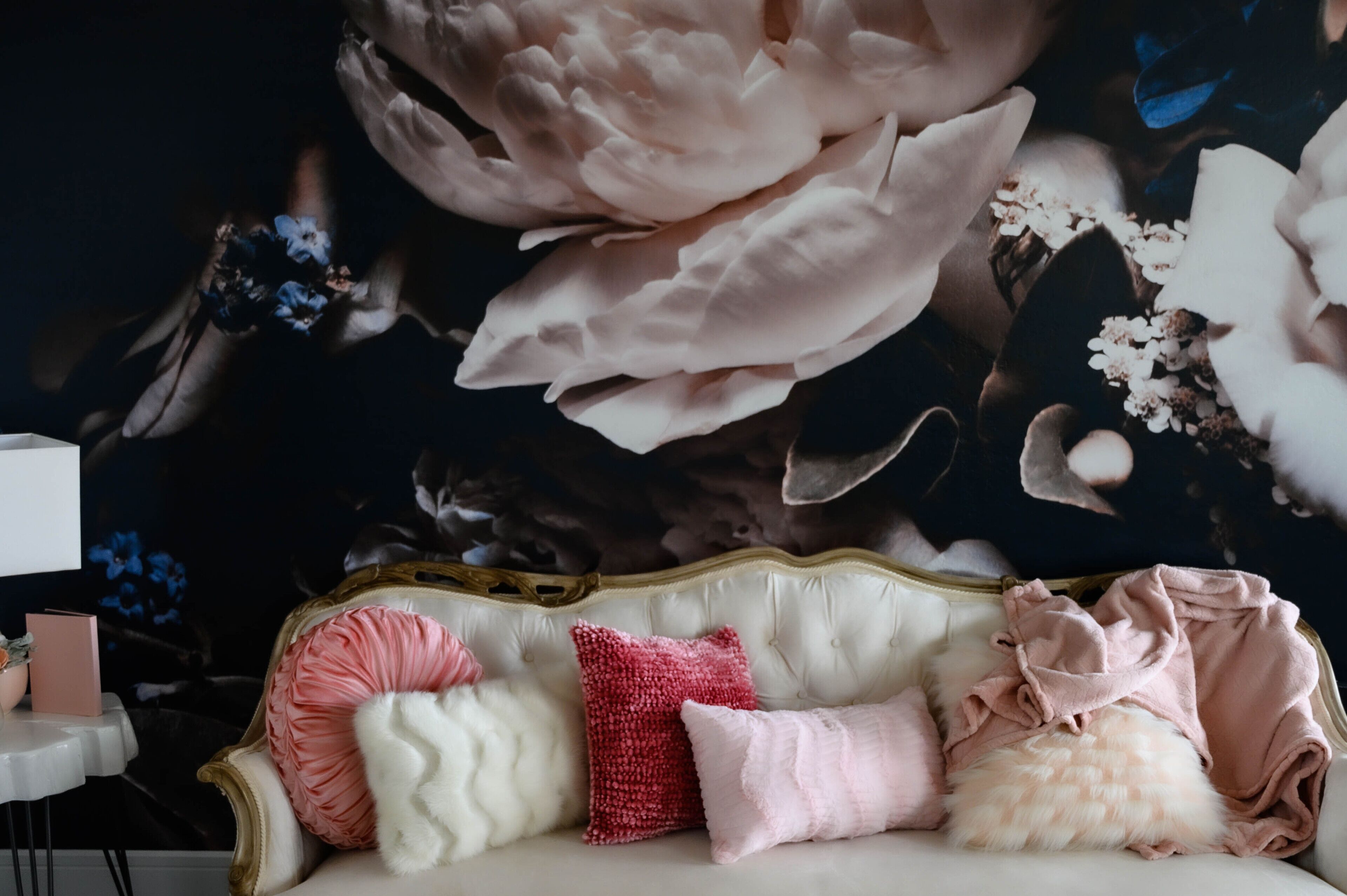 Una habitación con un amplio mural floral en la pared, un sofá blanco con varios cojines y una pequeña mesa redonda con un libro rosa y un jarrón.