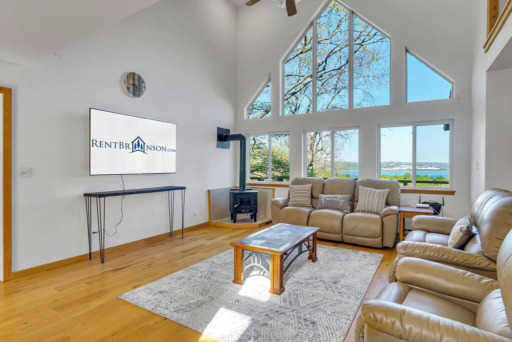 A spacious living room with a high ceiling, large windows, a fireplace, and a television mounted on the wall.