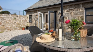 Un bâtiment en pierre avec une porte en verre, des meubles en osier et une table ornée d’un vase de fleurs.