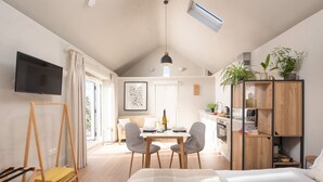 Une pièce moderne et bien éclairée, comprenant un lit, une table à manger avec des chaises, un téléviseur fixé au mur et un meuble en bois agrémenté de plantes.
