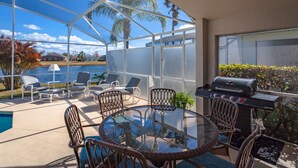 Une terrasse couverte avec une table et des chaises en verre, un barbecue et une vue sur un lac et des palmiers.