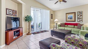Un salon comprenant un téléviseur, un ventilateur de plafond, un canapé vert, un pouf à motifs et une plante décorative.