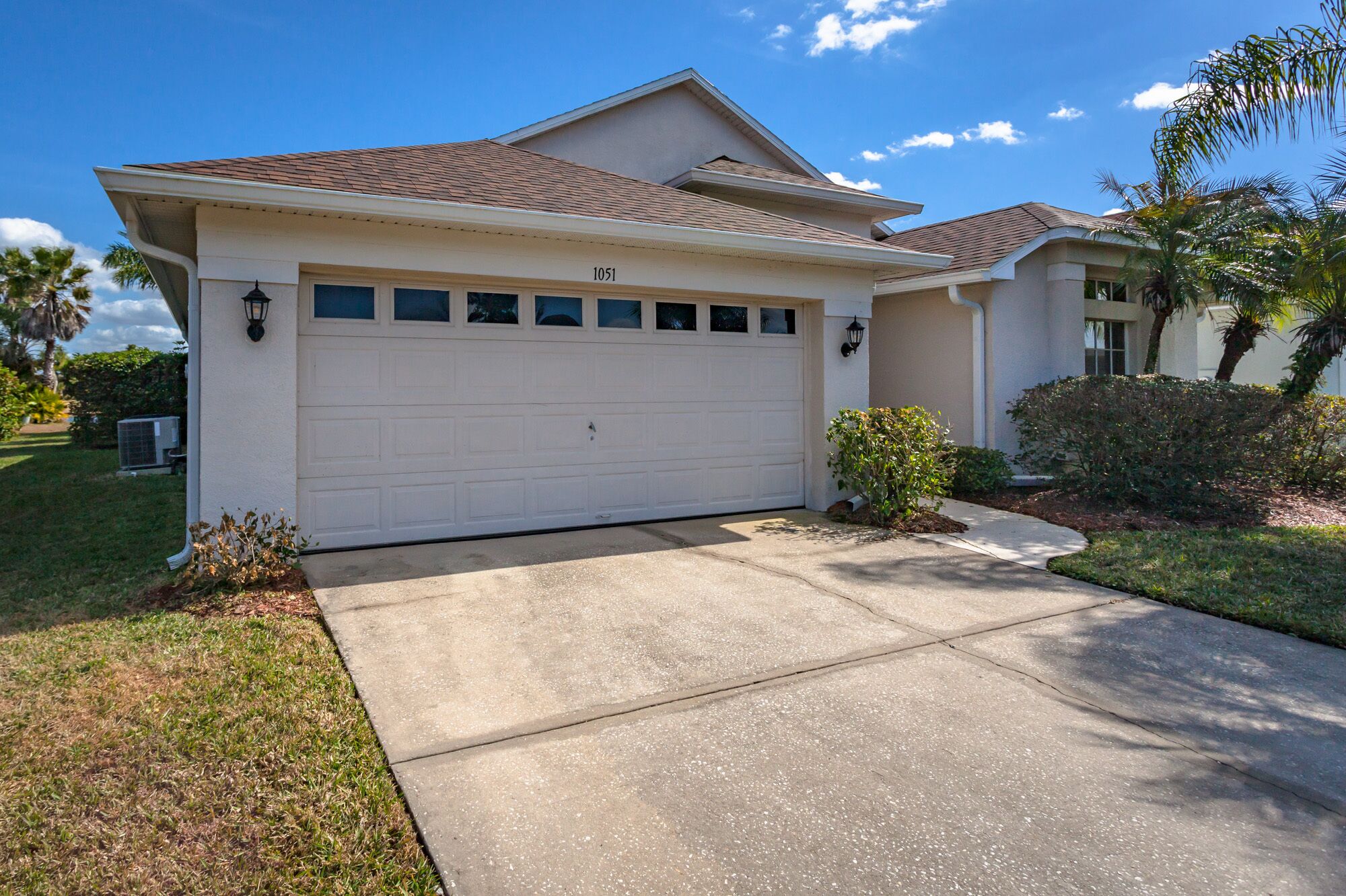 A single-story house with a white garage door, a small front yard with a bush, and a clear blue sky.