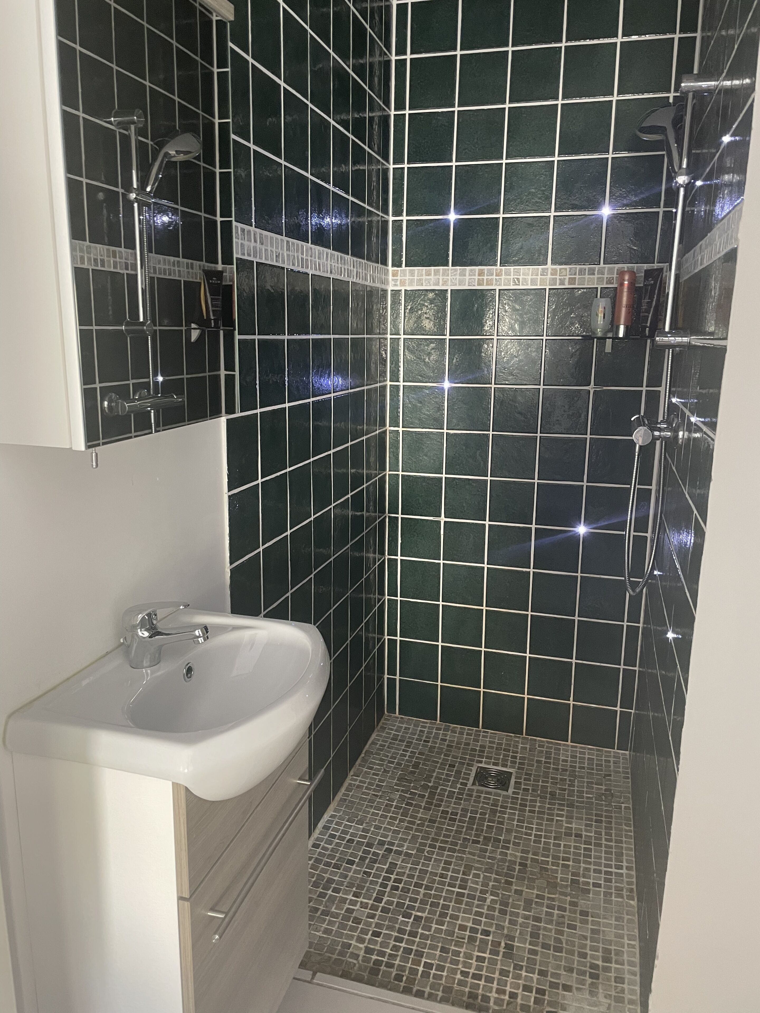 Un baño con lavamanos blanco, ducha con cabezal de ducha de mano y suelo de baldosas.