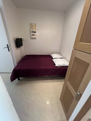 Ein kleines Zimmer mit einem Einzelbett, einer roten Bettdecke, weißen Kissen, einem an der Wand montierten Fernseher und einer Steckdose.