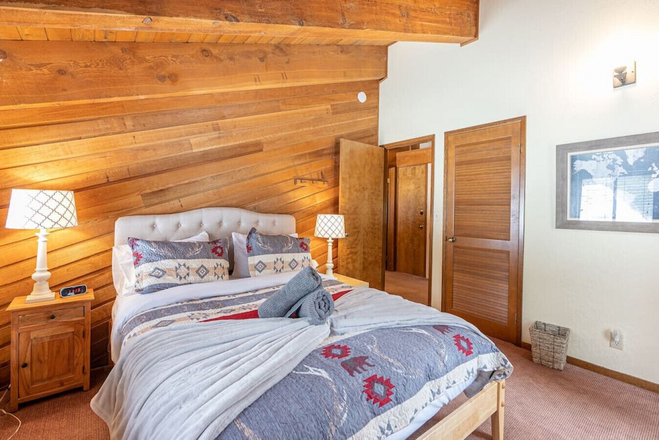 A bedroom with a wooden ceiling, a bed with patterned bedding, two bedside lamps, and a window with a view.