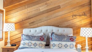 A bedroom with a wooden ceiling, a tufted headboard, two bedside tables with lamps, and patterned pillows.