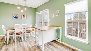 Un comedor con mesa de madera y sillas blancas, una ventana con vista a una terraza, y decoración mural.