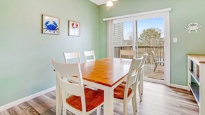 Un comedor con una mesa de madera y sillas blancas, una puerta corrediza de vidrio a un patio exterior, y dos cuadros en la pared.