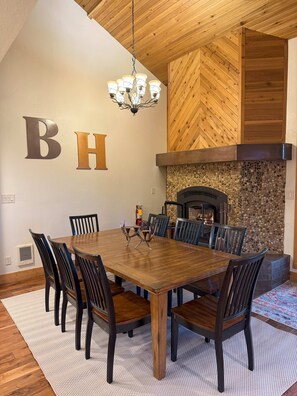Un espace repas avec une grande table en bois, des chaises sombres, un lustre et une cheminée avec un mur en pierre.