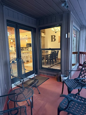 A covered outdoor seating area with metal chairs and a table, a wooden deck, and a view into a kitchen area with wooden cabinets and a chandelier.