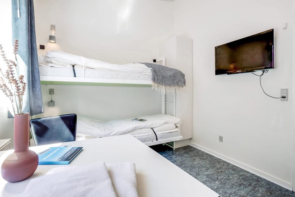 A bunk bed room with a TV mounted on the wall, a bedside table with a book and a vase, and a window with curtains.