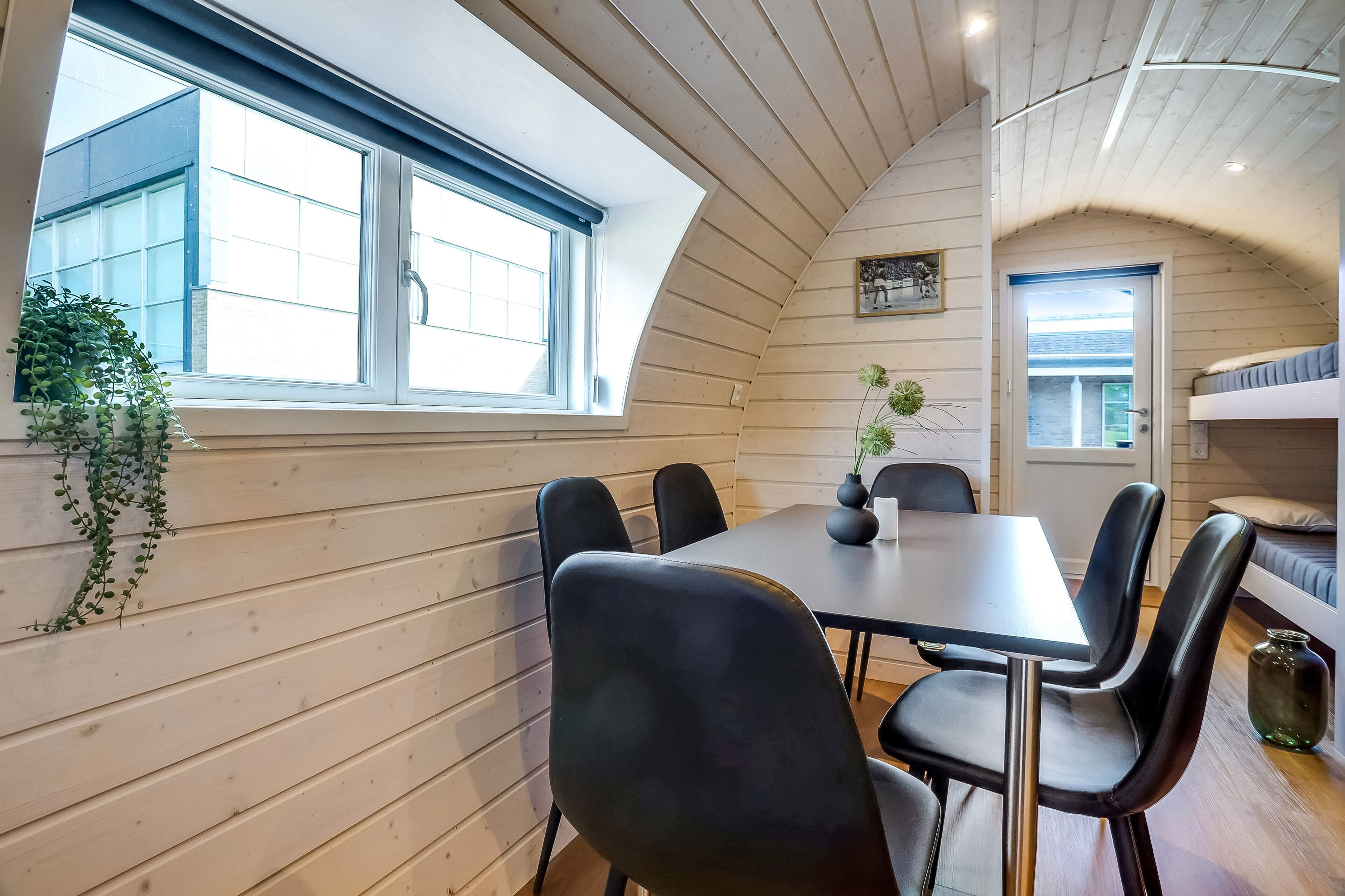 Een gezellige kamer met een houten inrichting, een eettafel met stoelen, een stapelbed en een raam met uitzicht op een gebouw.