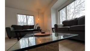 Ein modernes Wohnzimmer mit einem Glastisch, zwei dunklen Sofas und großen Fenstern mit Blick auf kahle Bäume.