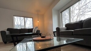 Una sala de estar moderna con una mesa de centro de vidrio, dos sofás oscuros y grandes ventanales con vistas a árboles desnudos.
