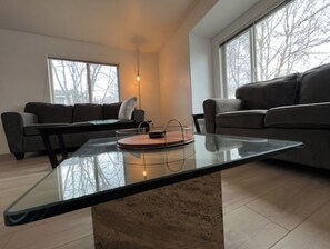 Una sala de estar moderna con una mesa de centro de vidrio, dos sofás oscuros y grandes ventanales con vistas a árboles desnudos.