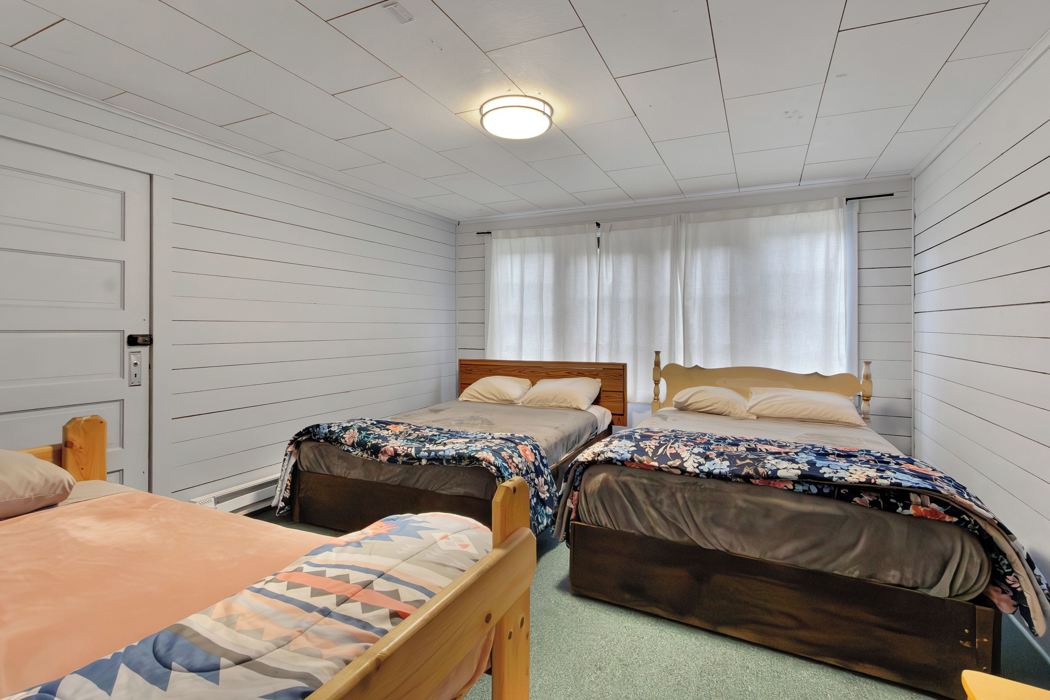 Une chambre avec deux lits, une tête de lit en bois, une fenêtre avec des rideaux et un luminaire au plafond.