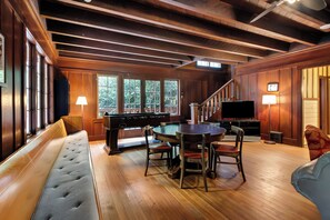 A wooden-floored room with a billiards table, a sofa, a dining table with chairs, and a television.