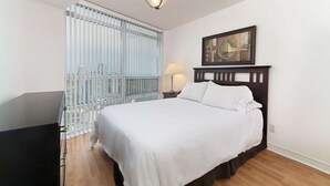 A hotel room with a bed, a nightstand with a lamp, a painting on the wall, and a view of the city through the window. - 2 Bedroom/2 Bathroom Suite (Scarborough)