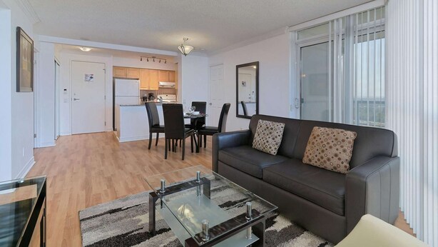 A modern living room with a glass coffee table, a dark sofa with patterned cushions, and a dining area with a kitchen in the background. - 2 Bedroom/2 Bathroom Suite (Scarborough)