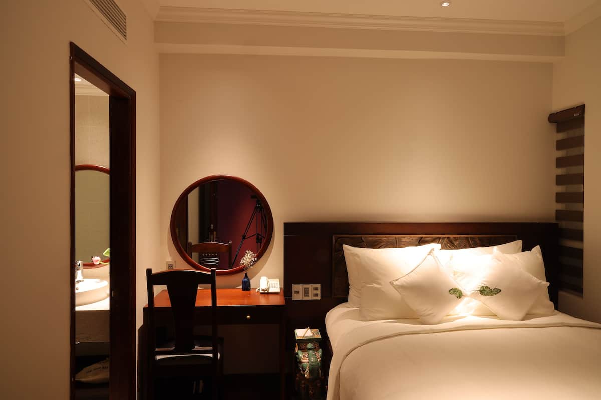 A hotel room with a bed, a wooden nightstand, a round mirror, a desk with a chair, and a small table with a vase.