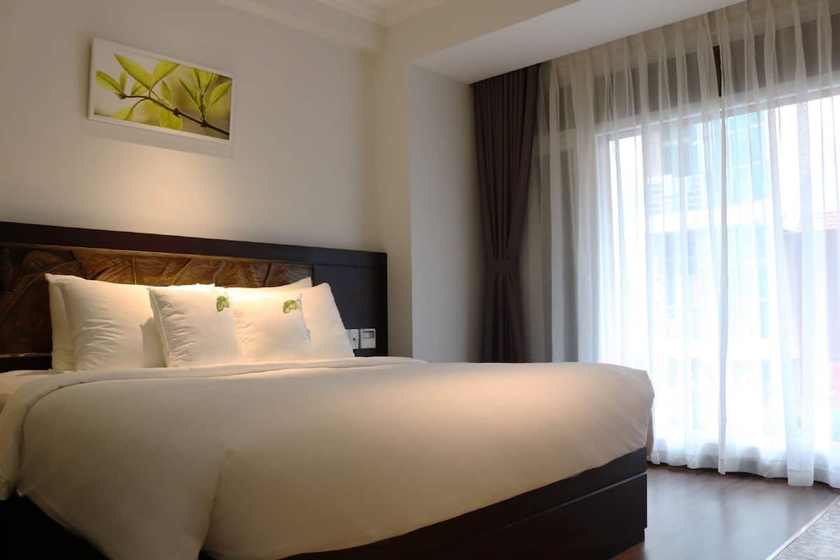 A large bed with white linens and a dark headboard, a wall-mounted plant, and sheer curtains on a window.