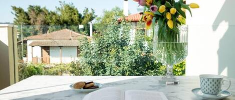 A table with an open book, a glass vase with flowers, a plate of cookies, and a cup on a balcony overlooking a house and greenery.