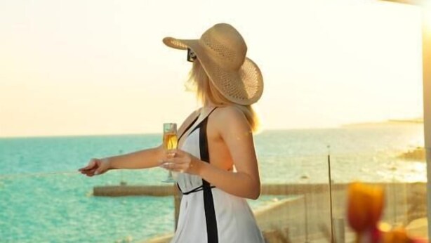 Una mujer con vestido blanco y sombrero de paja parada junto a una ventana con vista al océano.
