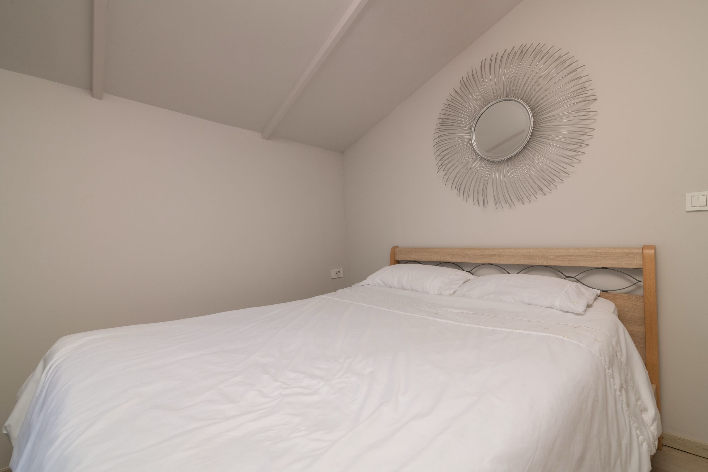 Ein ordentlich bezogenes Bett in einem Zimmer mit einem Holz-Kopfteil, einem Wandspiegel und einem Lichtschalter.