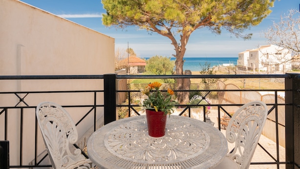 A balcony with a round table, two chairs, a potted plant, and a view of the sea and buildings. - Sweet Cottage Retreat (Zakynthos)