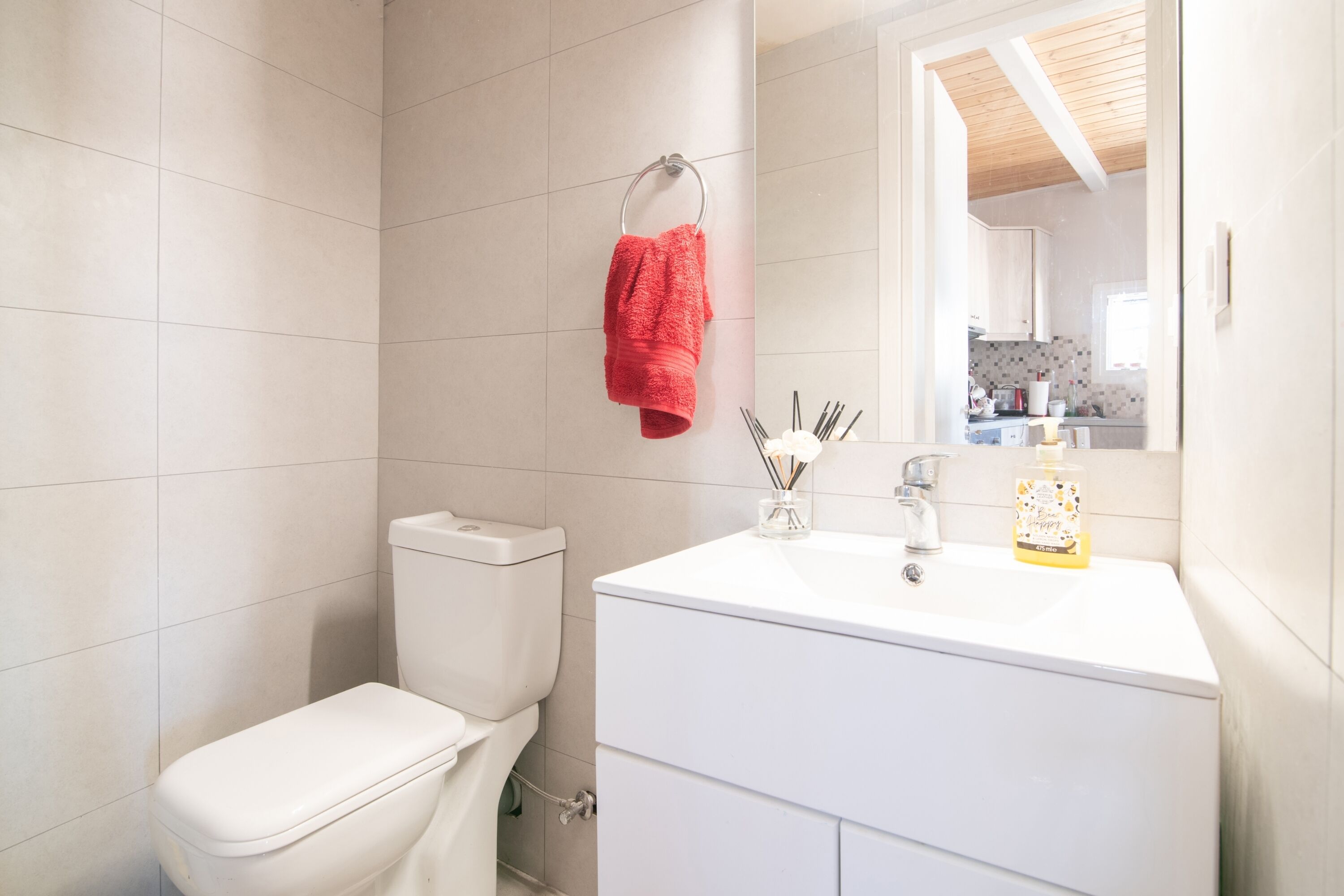 Un baño con un inodoro blanco, un lavabo blanco con espejo y una toalla roja colgada en un aro metálico.