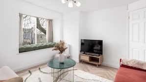 Ein Wohnzimmer mit Fernseher, einem Tisch mit einer Vase, einem Fenster mit Blick auf Gebäude und einer Tür.