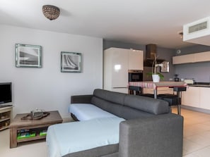 Un salon moderne avec un canapé gris, un téléviseur et une table basse. On aperçoit une cuisine à l’arrière-plan, avec une table à manger et des chaises.