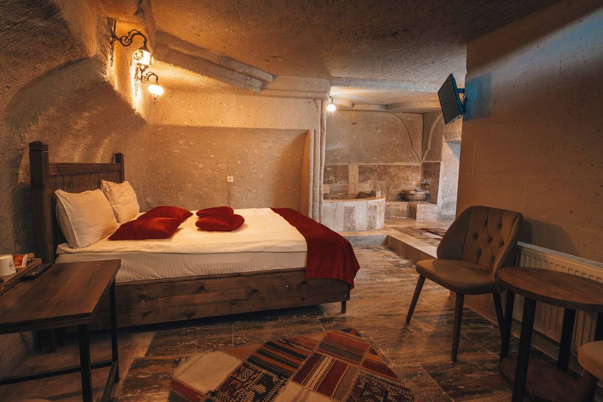 A rustic bedroom with a wooden bed, a small table, a chair, and a unique stone wall.