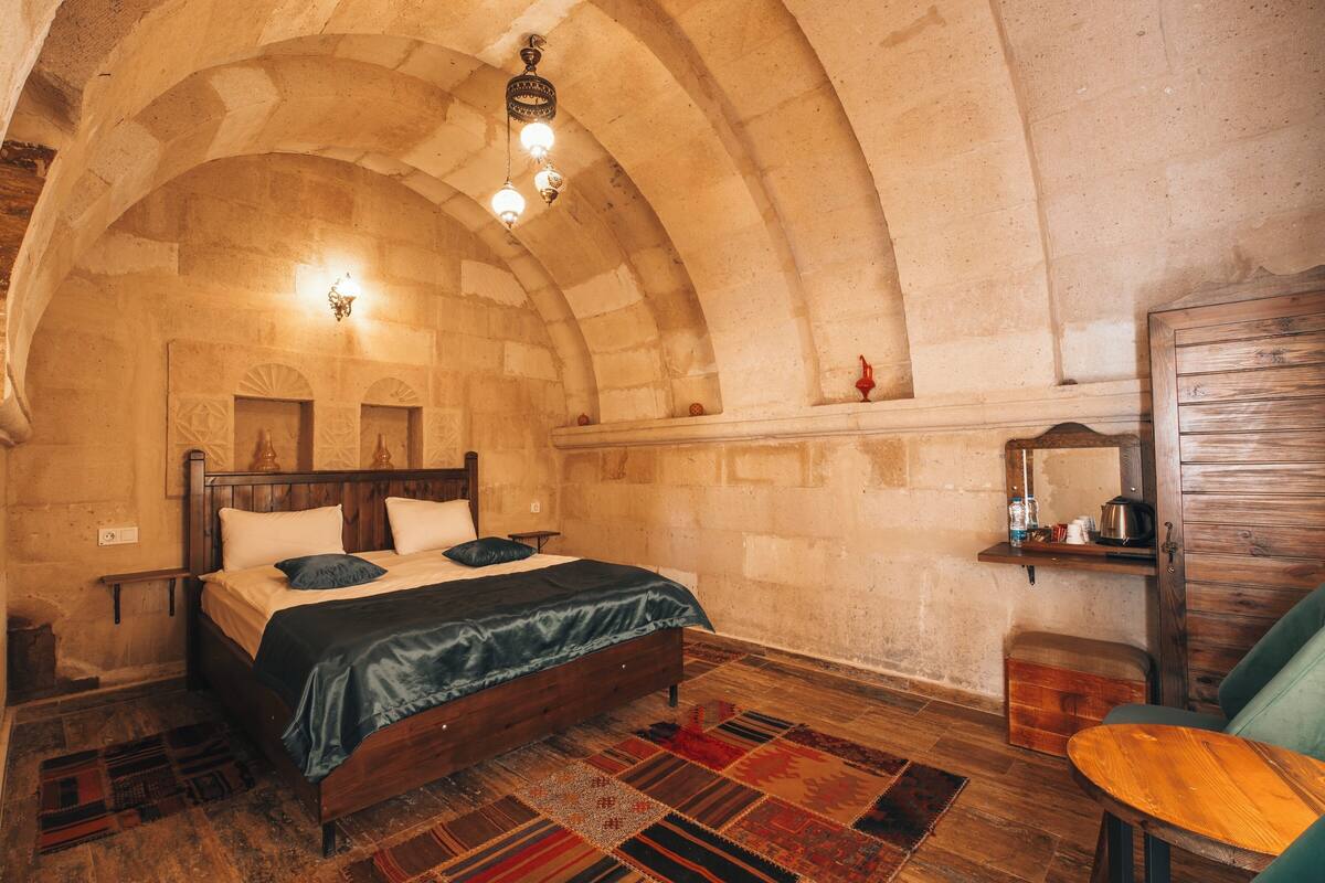 A room with a wooden bed, a small table, and a bench. The room has a stone wall and a wooden ceiling.