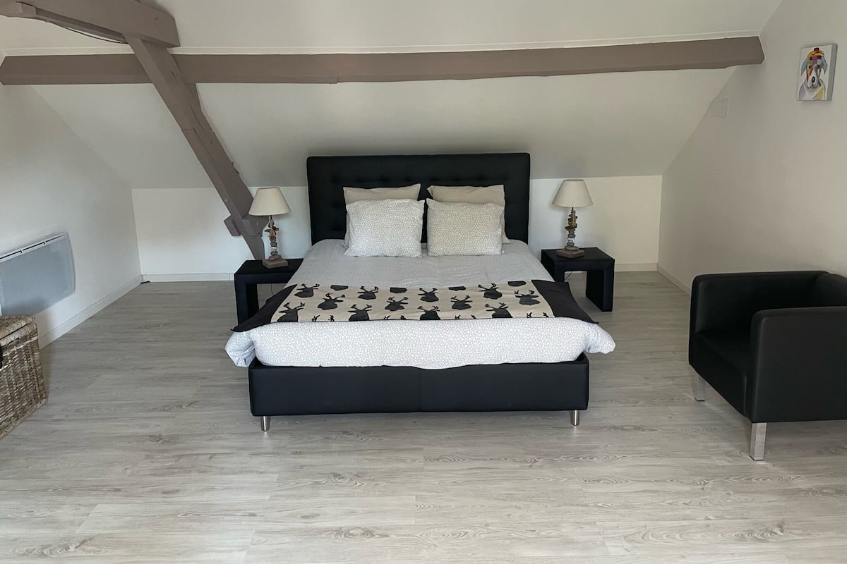A modern bedroom with a large bed, two bedside tables, a black chair, and a wooden ceiling beam.