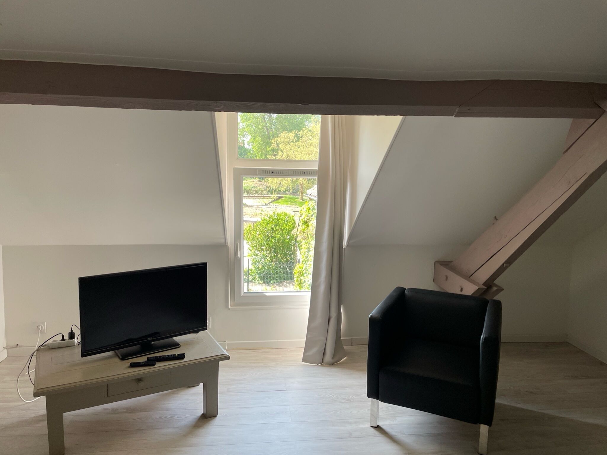 Une pièce avec une télévision posée sur une table blanche, une chaise noire et une fenêtre donnant sur un paysage verdoyant.