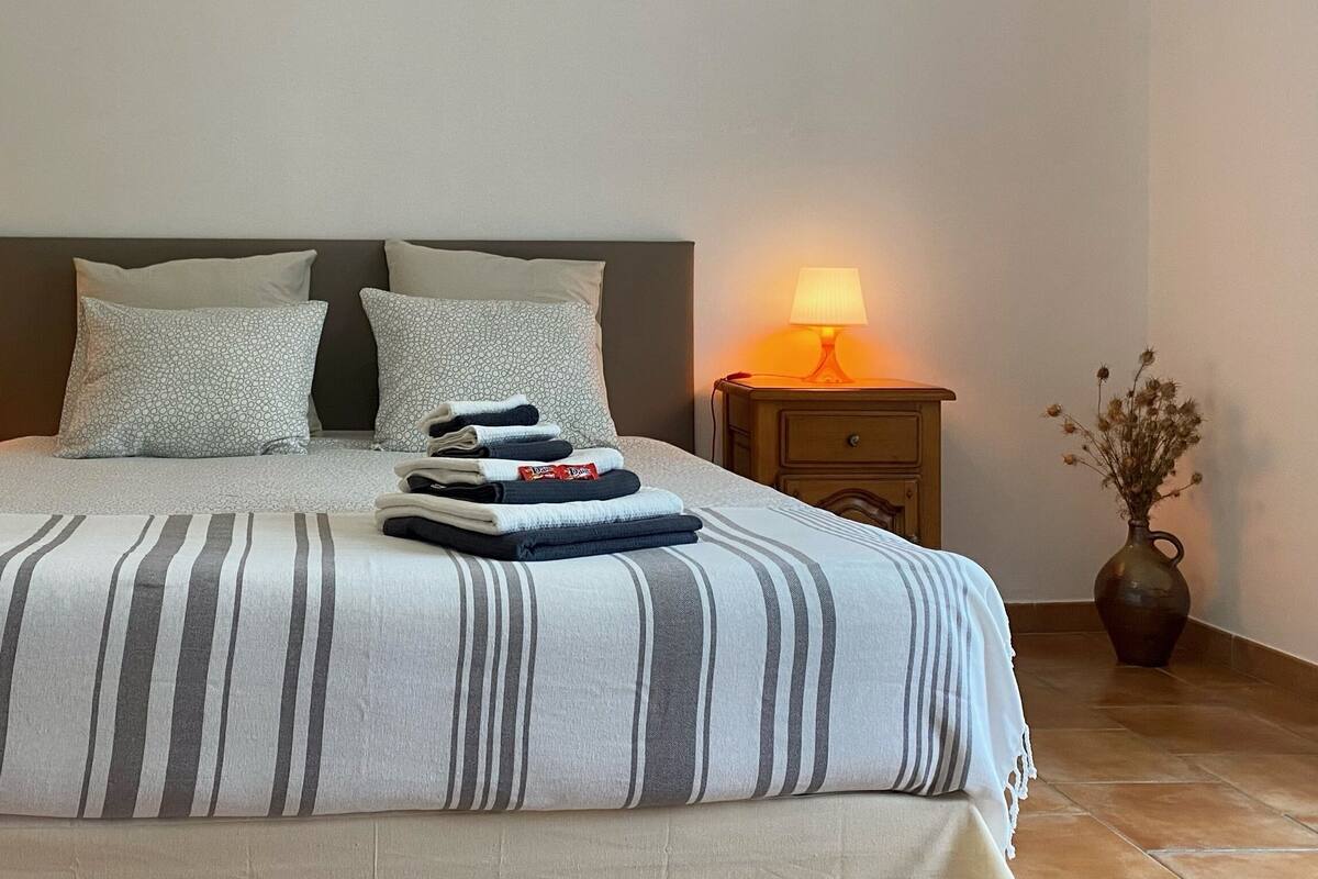 A neatly made bed with striped bedding, two pillows, and a stack of folded towels on the headboard. There are two bedside tables with lamps, and a vase with dried flowers on the floor beside the bed.