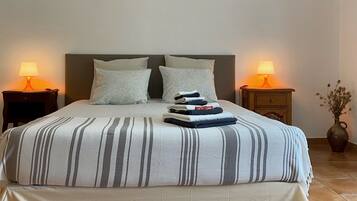 A neatly made bed with striped bedding, two pillows, and a stack of folded towels on the headboard. There are two bedside tables with lamps, and a vase with dried flowers on the floor beside the bed.