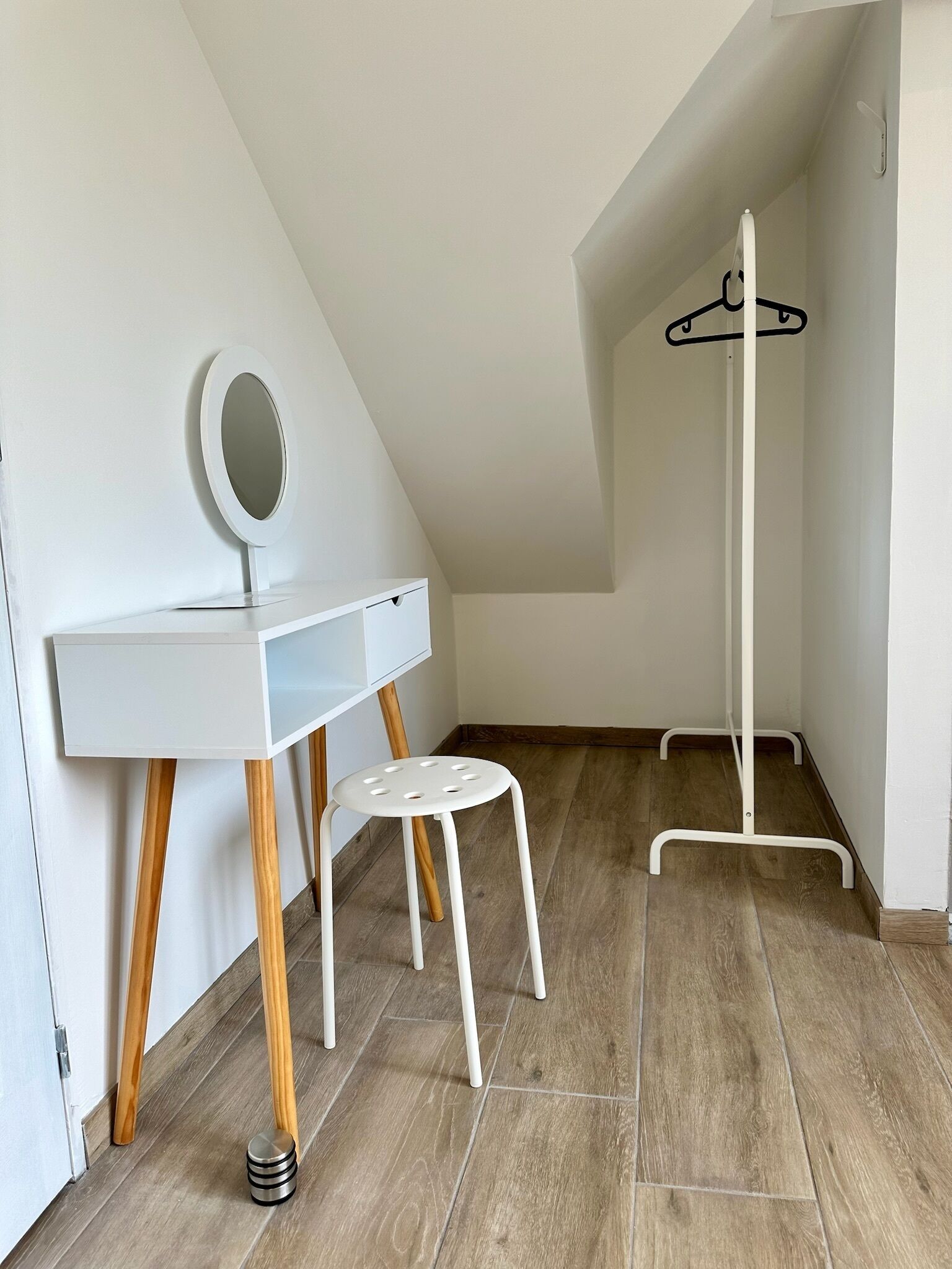A small, white room with a sloped ceiling, a white desk with a mirror, a wooden stool, and a coat rack.