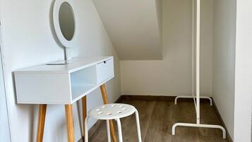 Una habitación pequeña y blanca con techo inclinado, un escritorio blanco con espejo, un taburete de madera y un perchero.