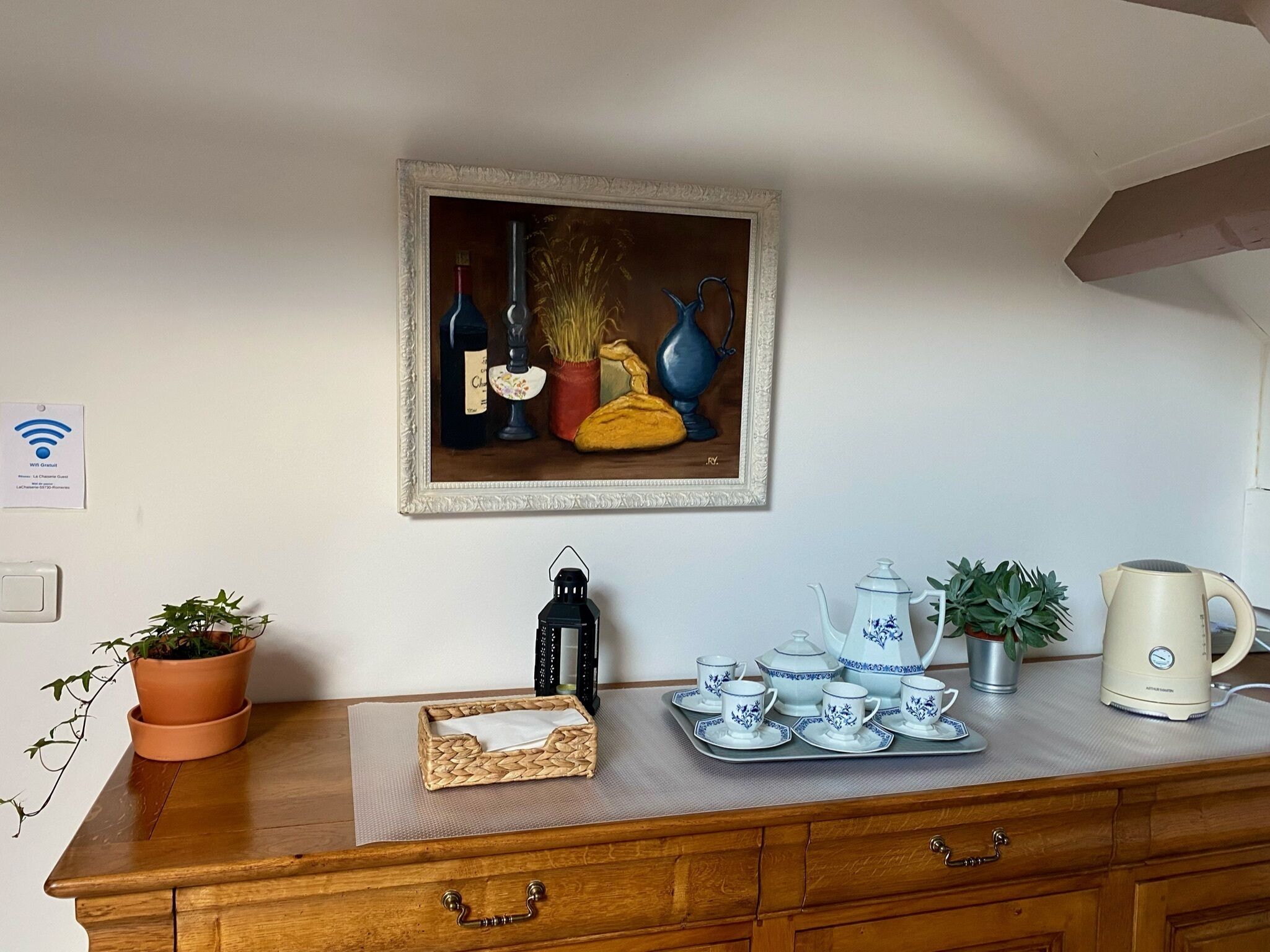 A kitchen counter with a framed painting, a kettle, a tray with teacups, and a potted plant.