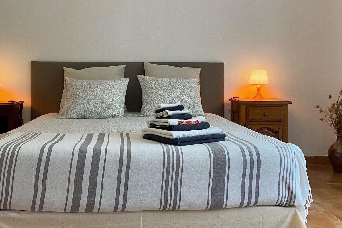 A neatly made bed with striped bedding, two bedside tables with lamps, and a vase with dried flowers on the floor.