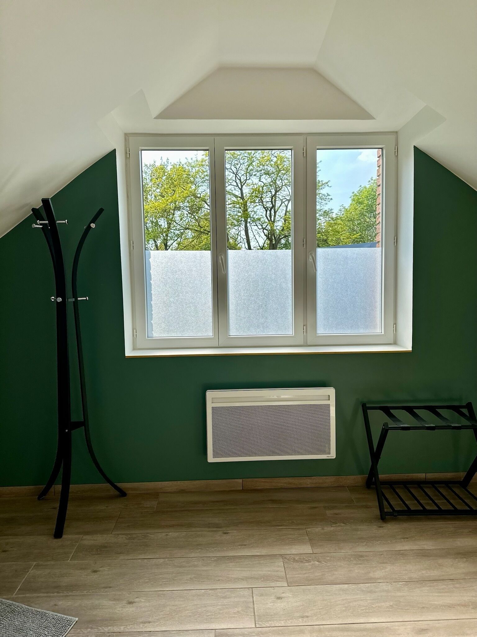 Una habitación con techo inclinado, una ventana con vidrio esmerilado, un radiador y una estantería de metal negro.