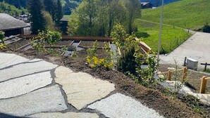 A mountainous landscape with a clear sky, a flag, and a stone pathway leading through a garden area. - Untere Bühlstrasse 10 - EG by Interhome (Lenk)