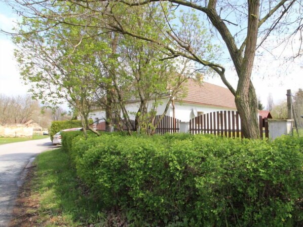 A white building with a red roof, surrounded by a wooden fence and dense green hedges.